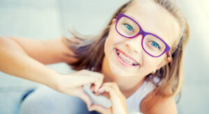 Smiling little girl with dental braces and glasses showing heart with hands